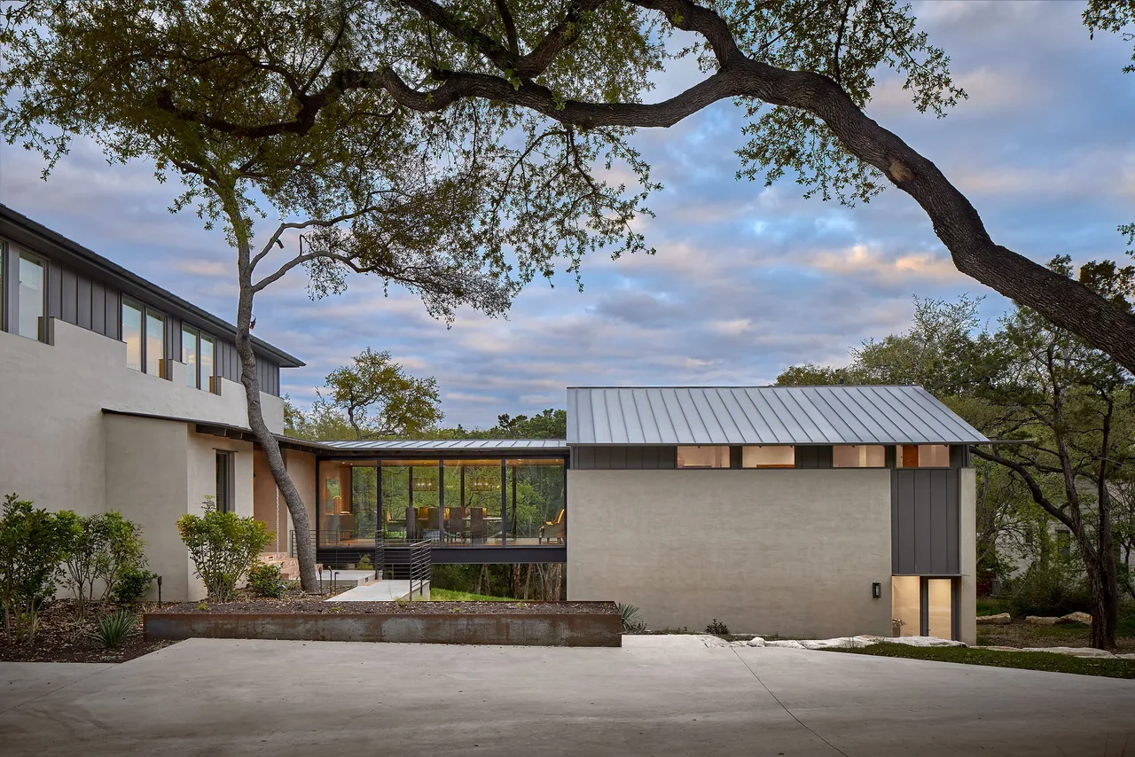 A modern house with a metal roof features a glass bridge connecting two sections, surrounded by trees at dusk.