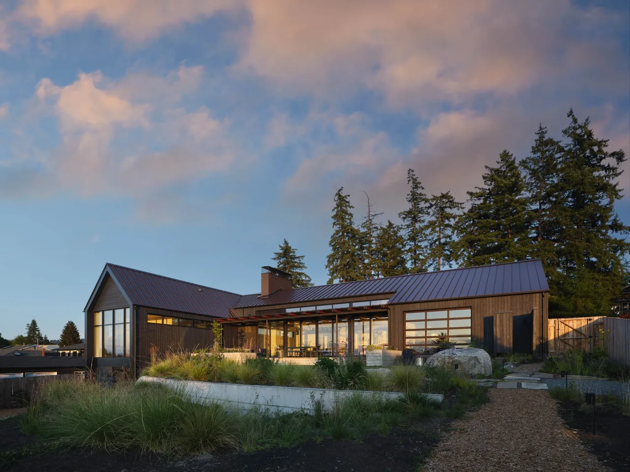 Modern house exterior with wood siding, large windows, and a brown metal gable roof at sunset under a partly cloudy sky.