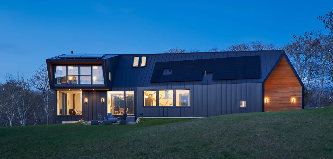 Modern black metal-sided house with large windows and solar panels at dusk on a green lawn.