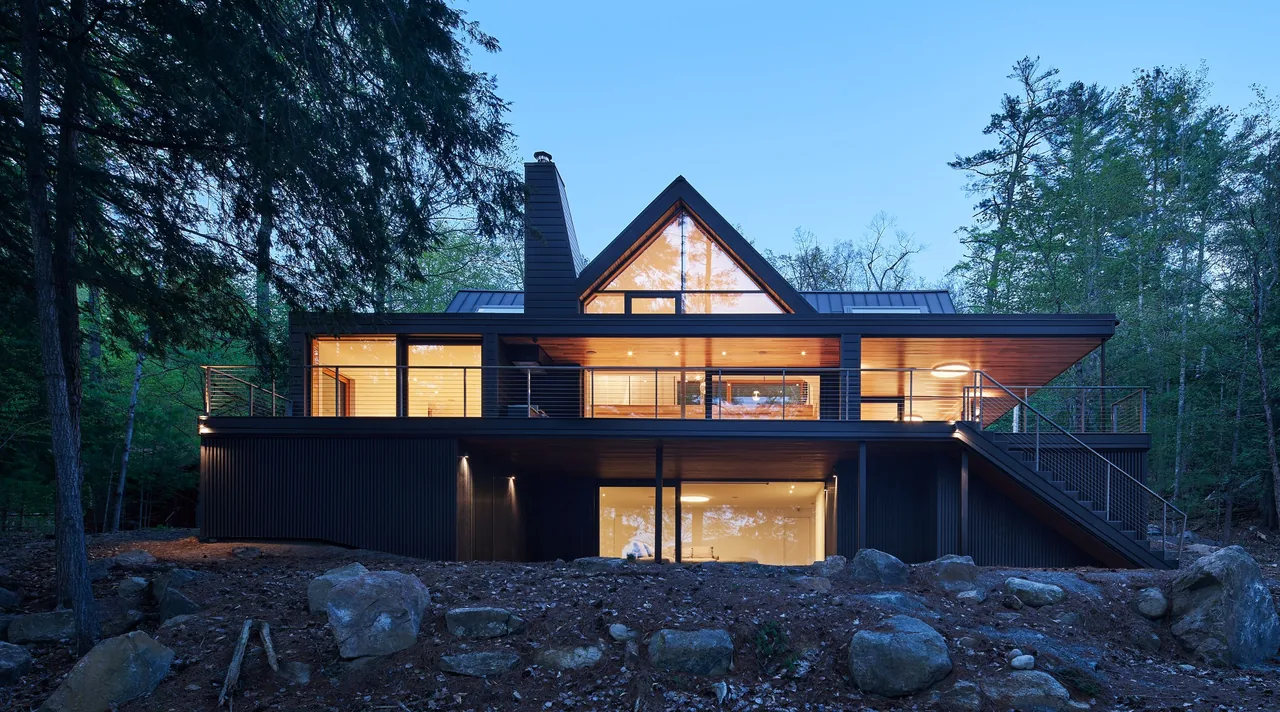 Exterior view of a modern house at dusk with large glass windows and a metal roof surrounded by trees.