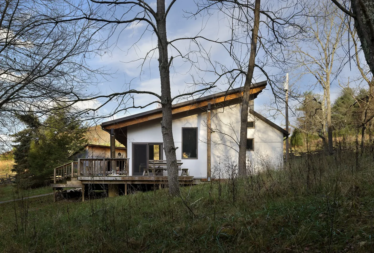 Side view of a white house with a raised wooden deck and a shed roof surrounded by leafless trees and grass.