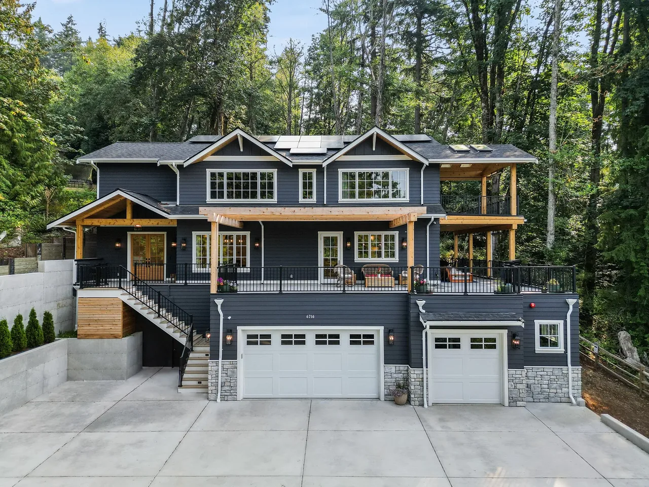 Two-story dark blue house with white trim, stone foundation, multiple garages, and a large porch with wooden beams, set against a background