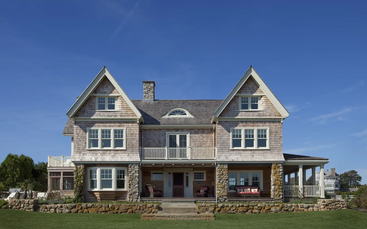Front view of a large two-story house with shingle siding, stone chimney and foundation, multiple gable roofs, and a porch with seating.