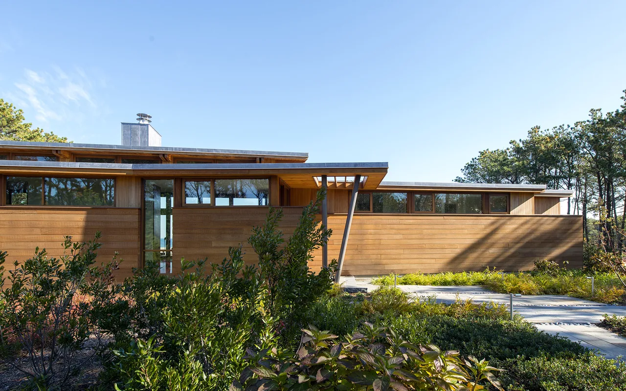 Single-story modern residence with horizontal wood siding and a flat roof surrounded by greenery under a clear blue sky.