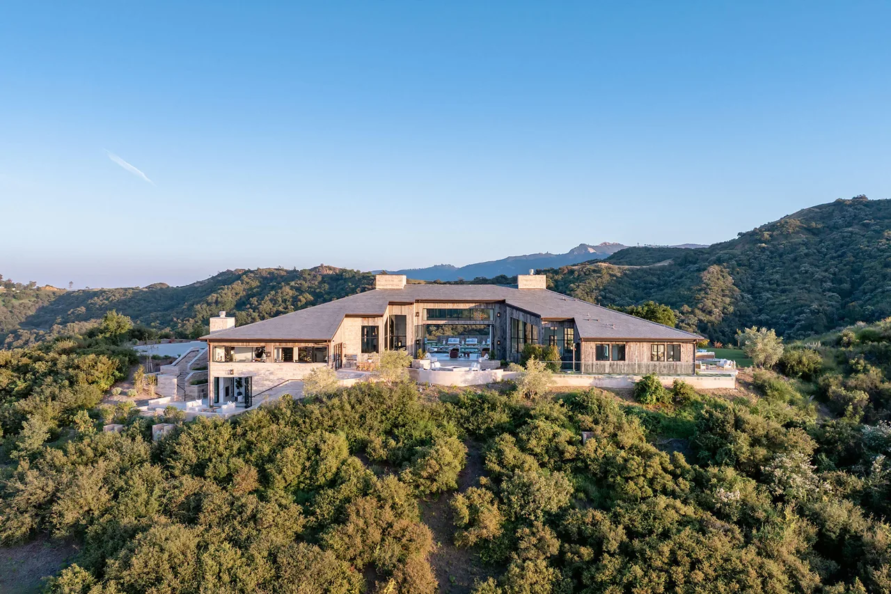 A modern house with large glass windows and courtyard set on a hill surrounded by green hills under a clear sky.