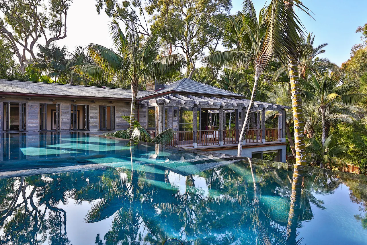 View of a modern house exterior with a large infinity edge pool surrounded by tropical trees.