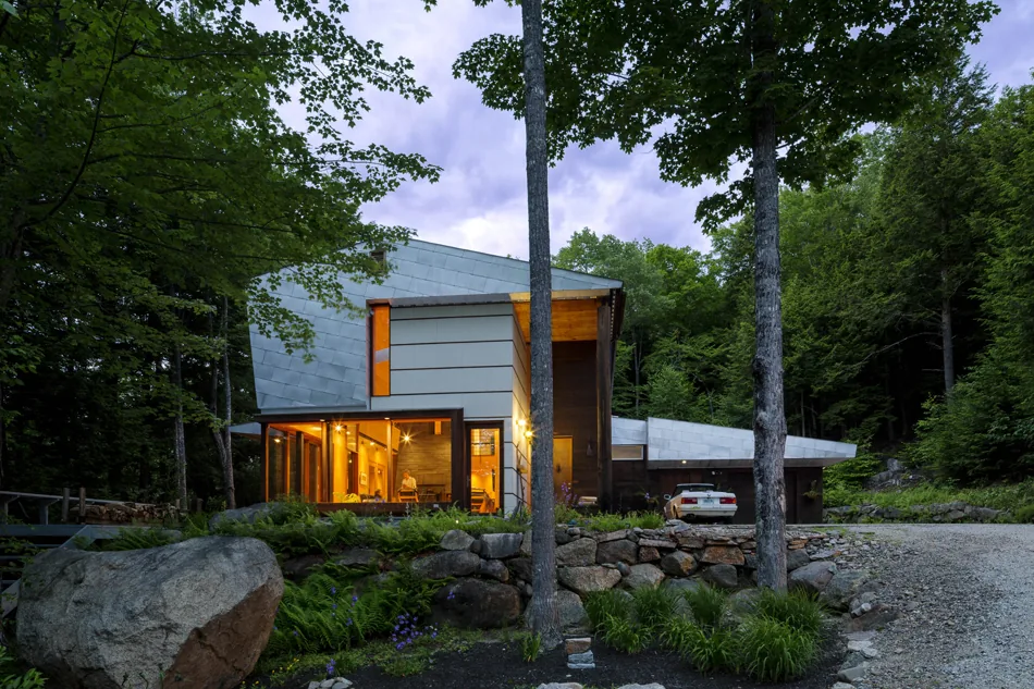 Modern two-story house in a forested mountain setting with large windows illuminated at twilight.