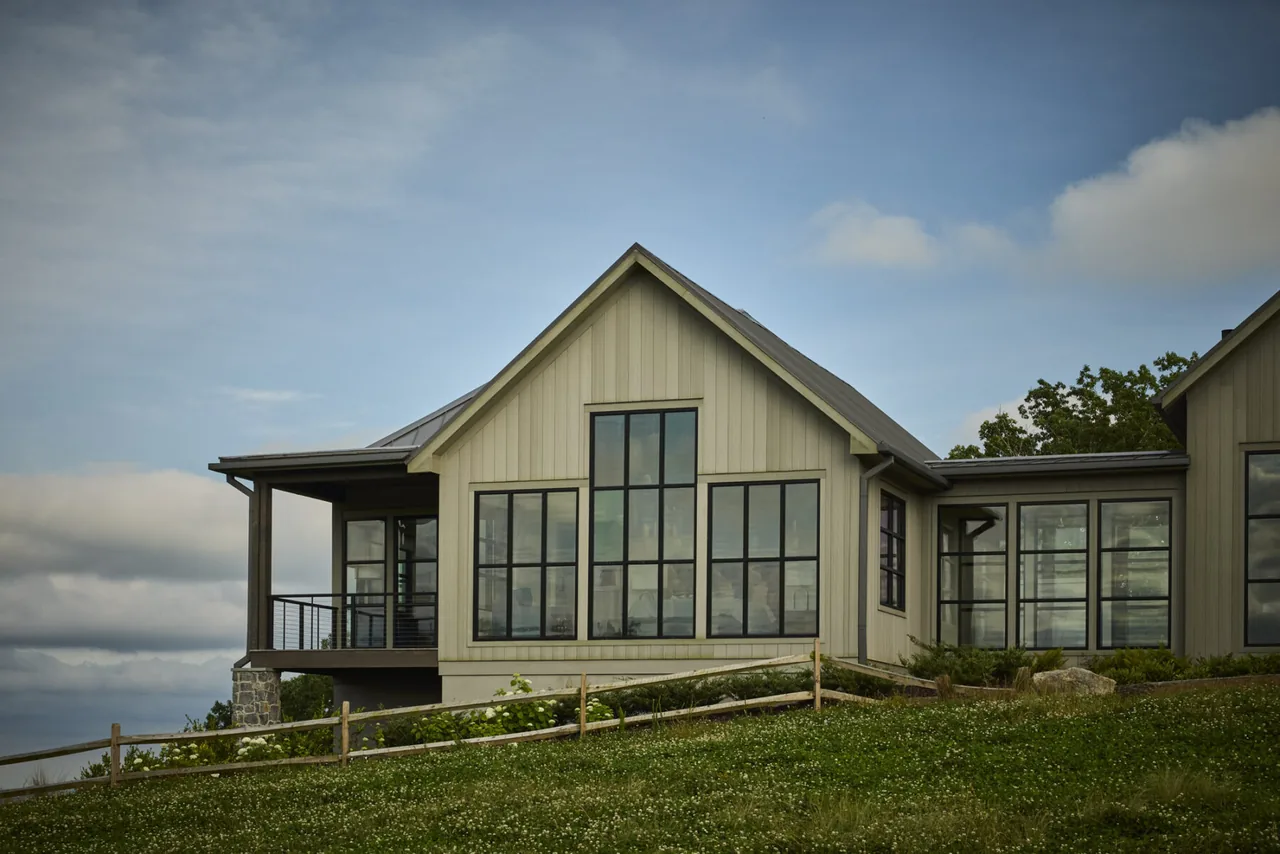 Exterior view of a modern farmhouse style house with large black-framed windows and a covered porch on a grassy hillside.