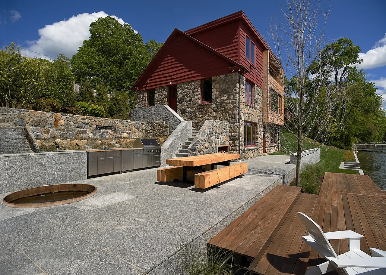 Stone and wood lakeside house with outdoor kitchen, picnic table, and dock under a blue sky with clouds.