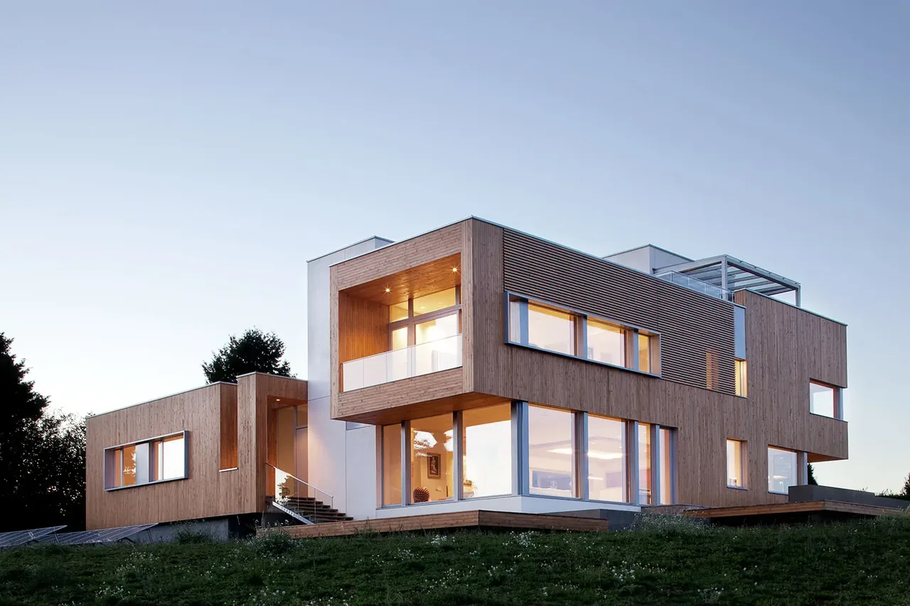 Modern two-story house made of wood with large windows and an overhanging upper level during dusk.