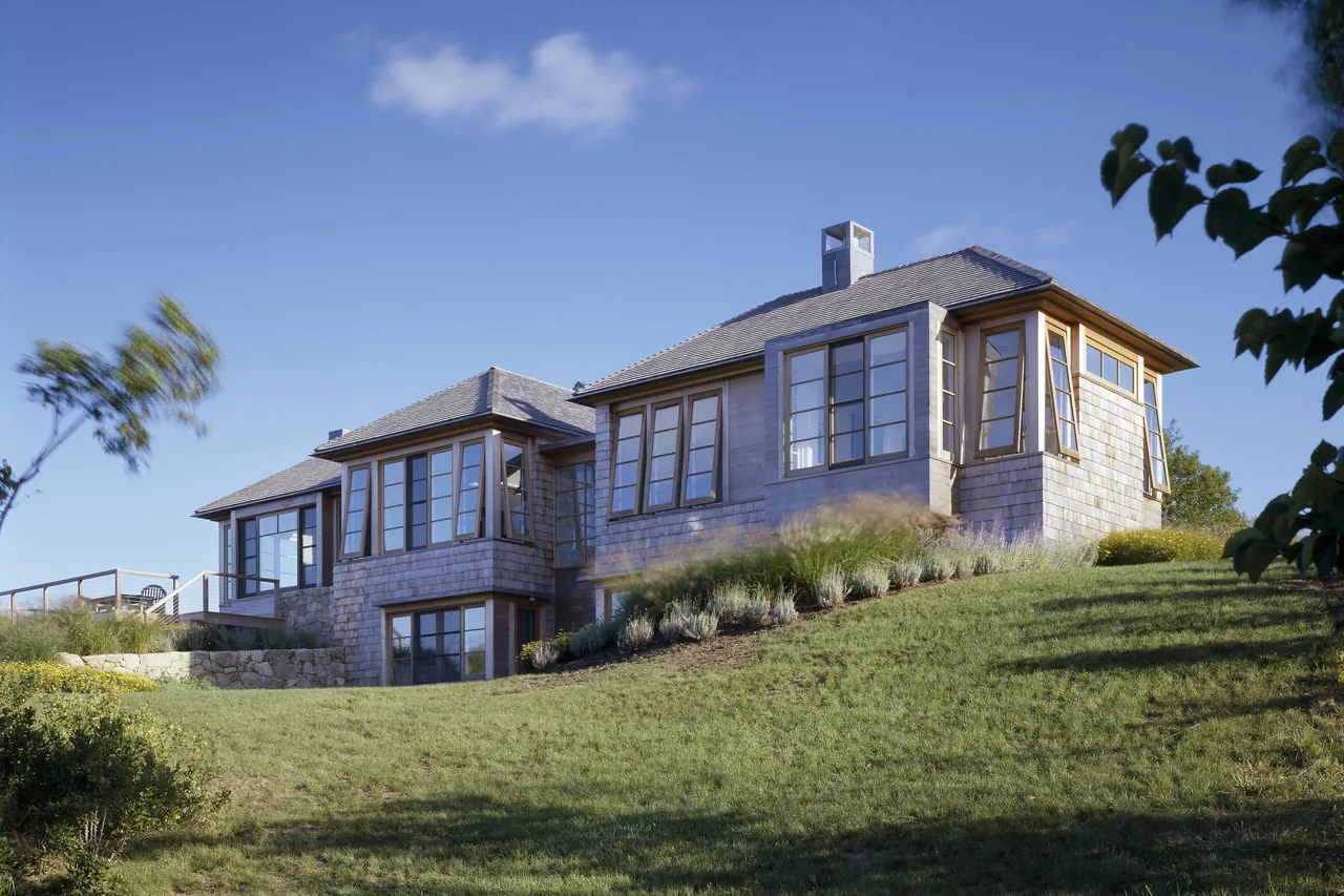 View of a shingle-style house on a grassy hill under a clear blue sky.