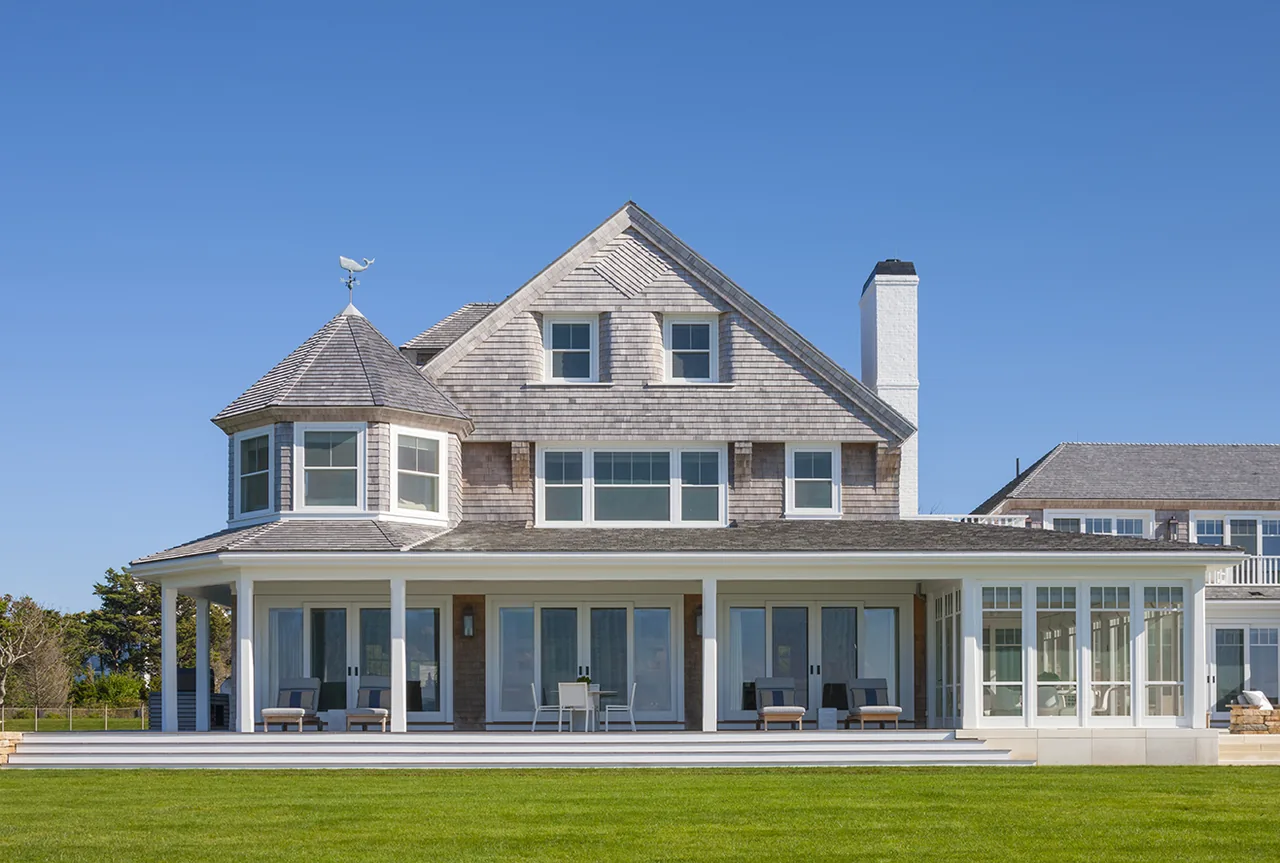 Exterior view of a traditional house with symmetrical shingles, multiple windows, and a large covered front porch with seating.