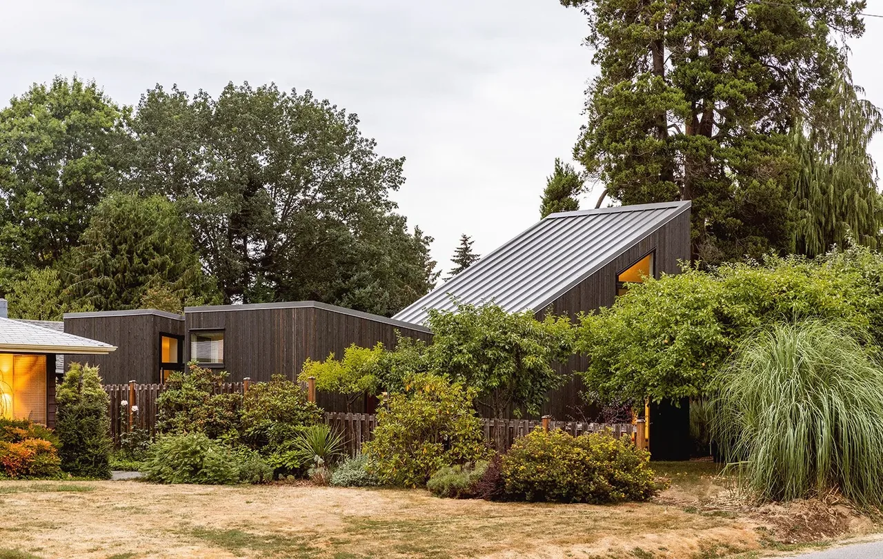 Exterior view of House O showing dark wood siding and a standing seam metal roof with surrounding trees and shrubs.