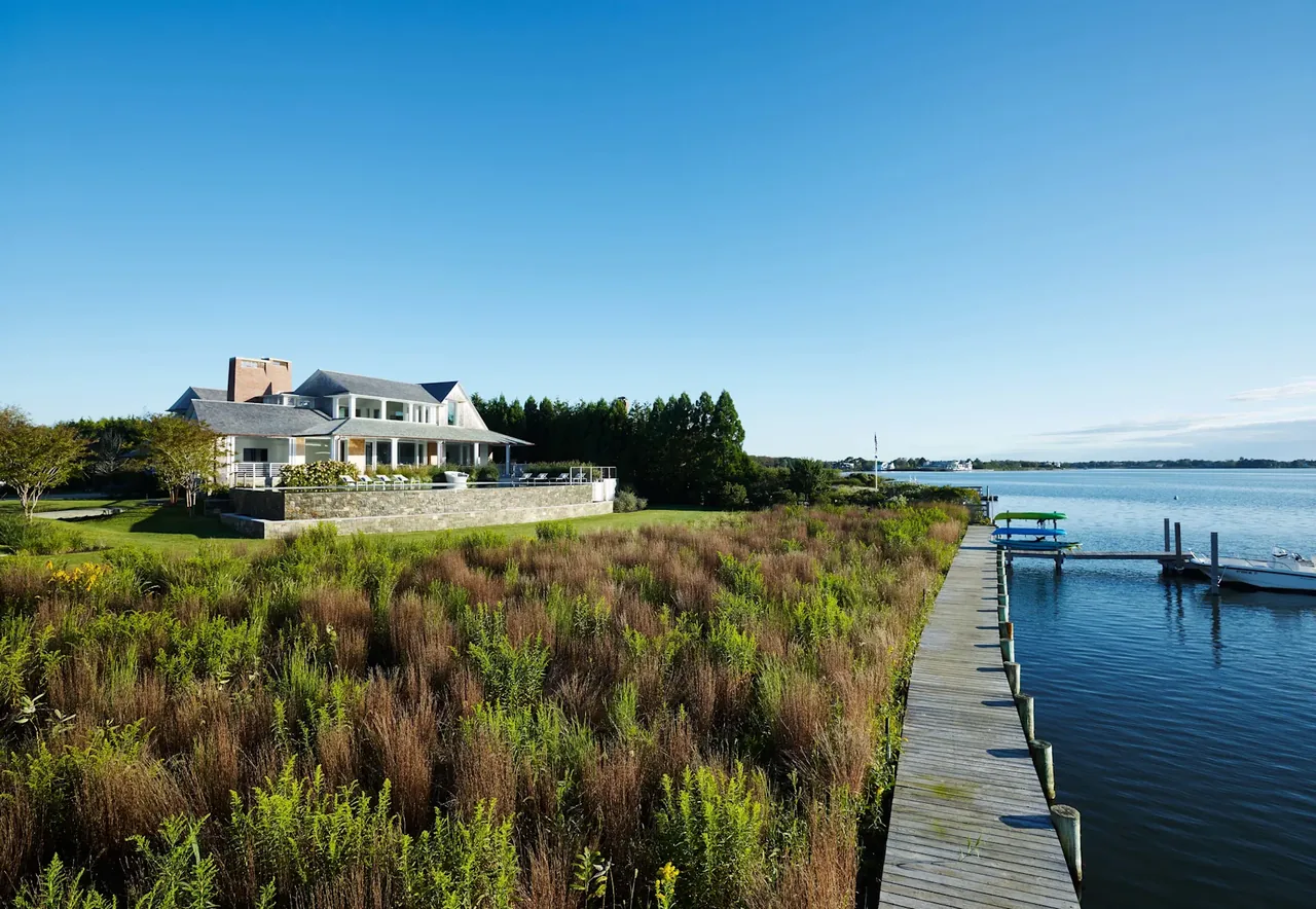 A large house on a waterfront property with a wooden dock extending into the water and grassy marshland in front.