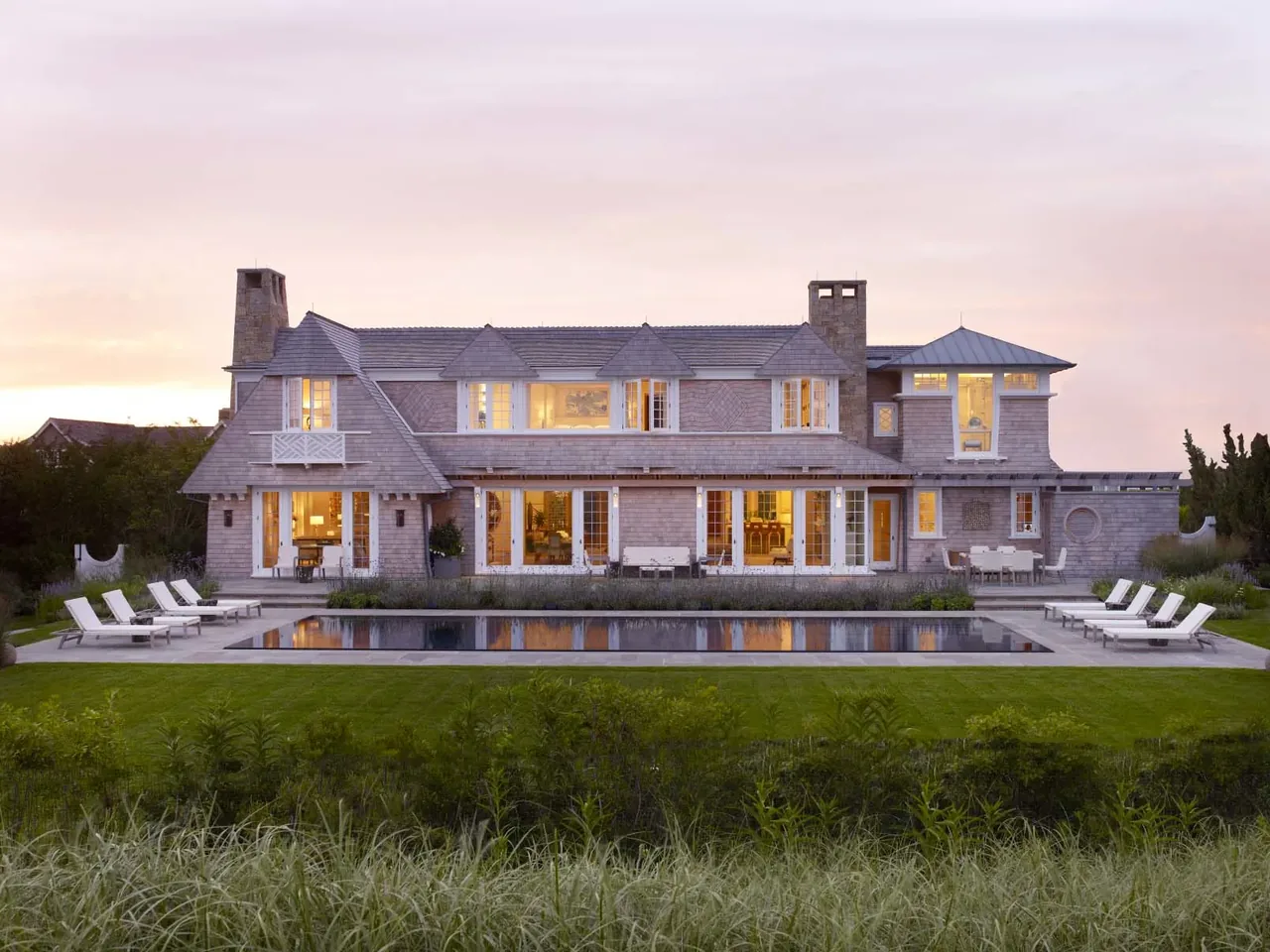 Shingle-style two-story beach house exterior with large windows and pool at sunset.