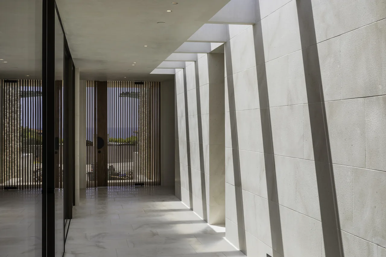 Sunlight casts vertical shadows through overhead beams onto a narrow hallway with light-colored tiled floors and walls.