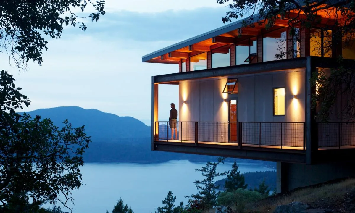 A modern cantilevered house at dusk with a person standing on the balcony overlooking a lake and mountains.