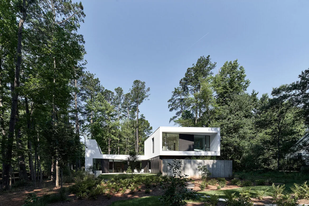 Modern two-story house with white and dark exterior surrounded by tall trees and greenery under clear blue sky.