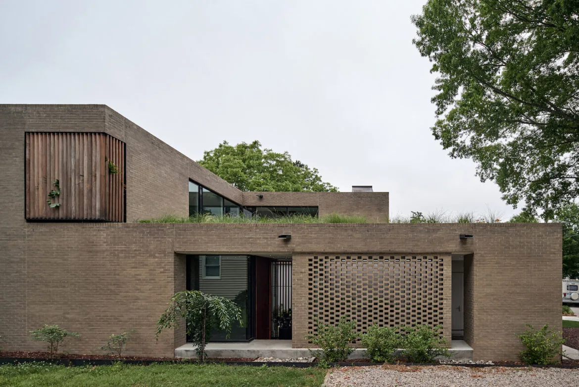 Exterior view of a modern two-story brown brick house with a green roof and wooden slat window detail.