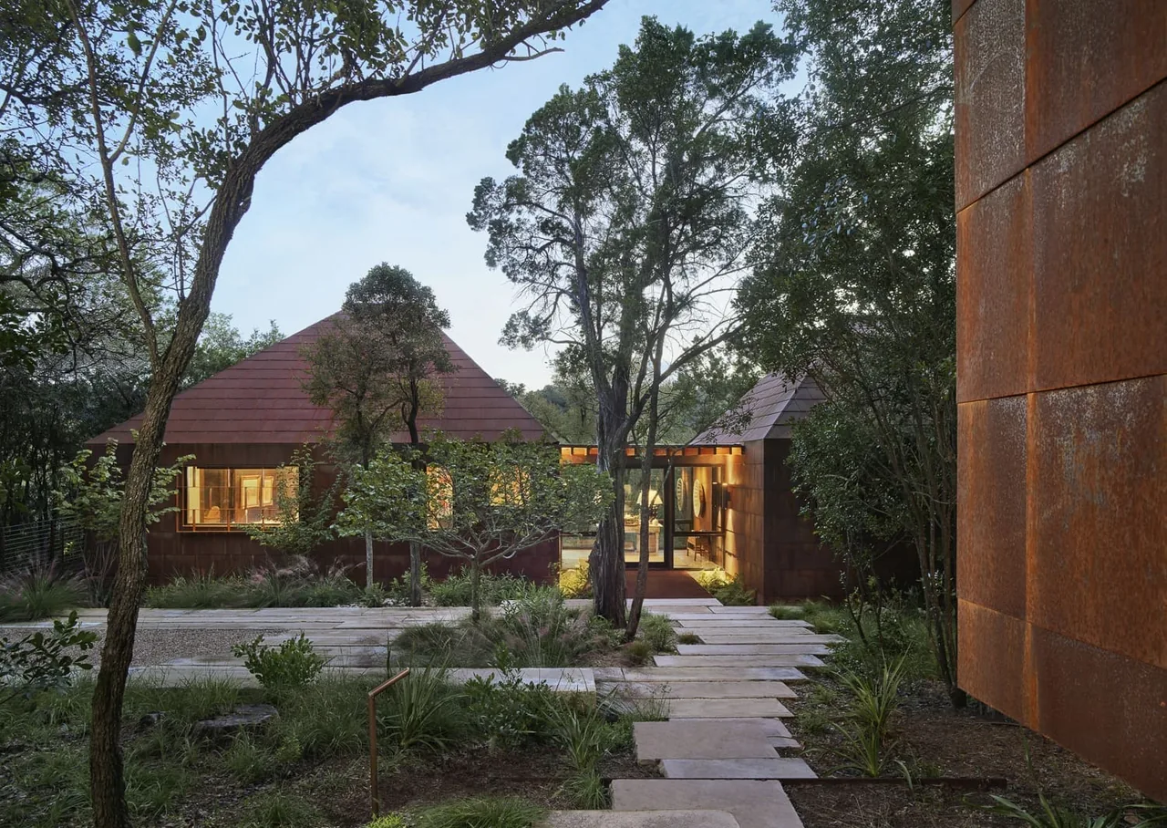 Exterior view of a modern rust-colored house with pyramid-style roofs surrounded by trees and greenery at dusk.