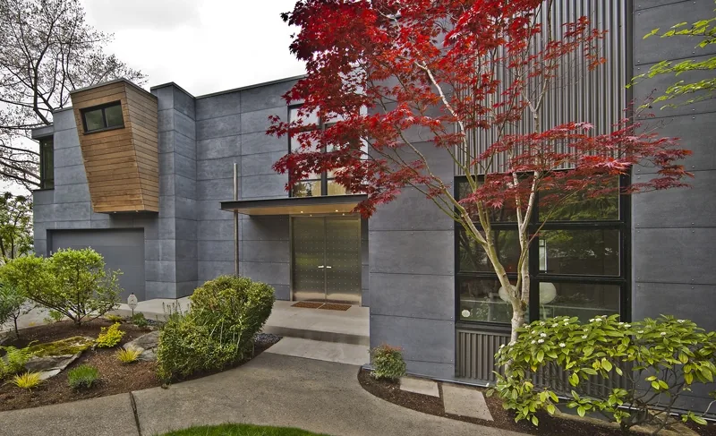 Modern house exterior with gray concrete walls, large windows, and a red-leafed tree in front.