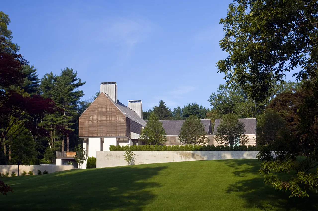 View of a modern house with sloped roofs and wood siding behind a concrete retaining wall on a large green lawn.