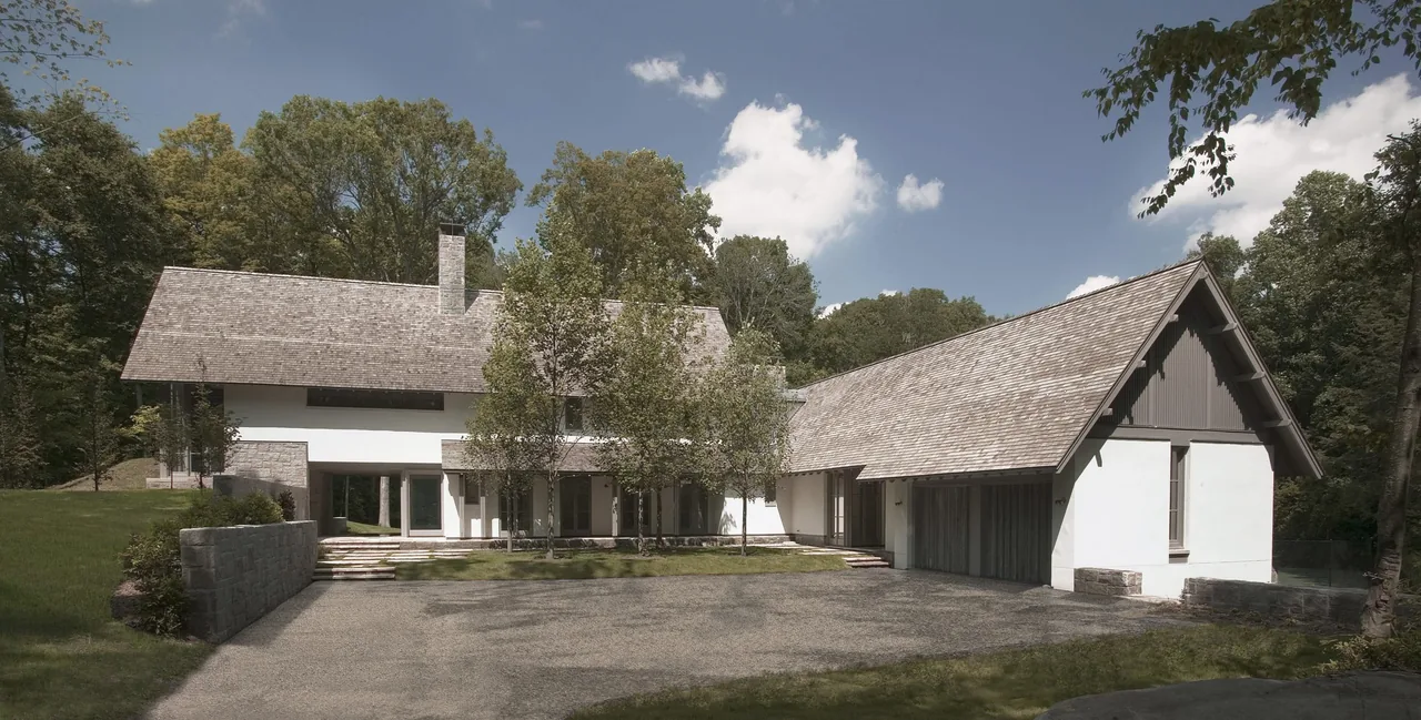 A residence with two large pitched roof sections and white walls set in a wooded area under a partly cloudy sky.
