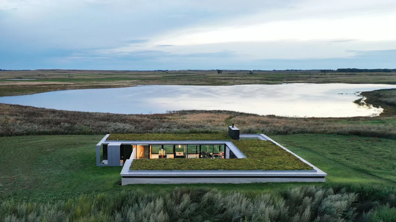 A modern single-story house with a green roof surrounds a central courtyard near a large body of water in a flat rural landscape.