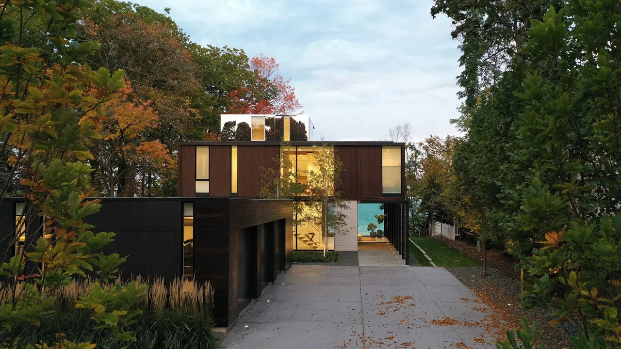 Modern two-story house with dark wood siding and large windows surrounded by trees with autumn foliage.