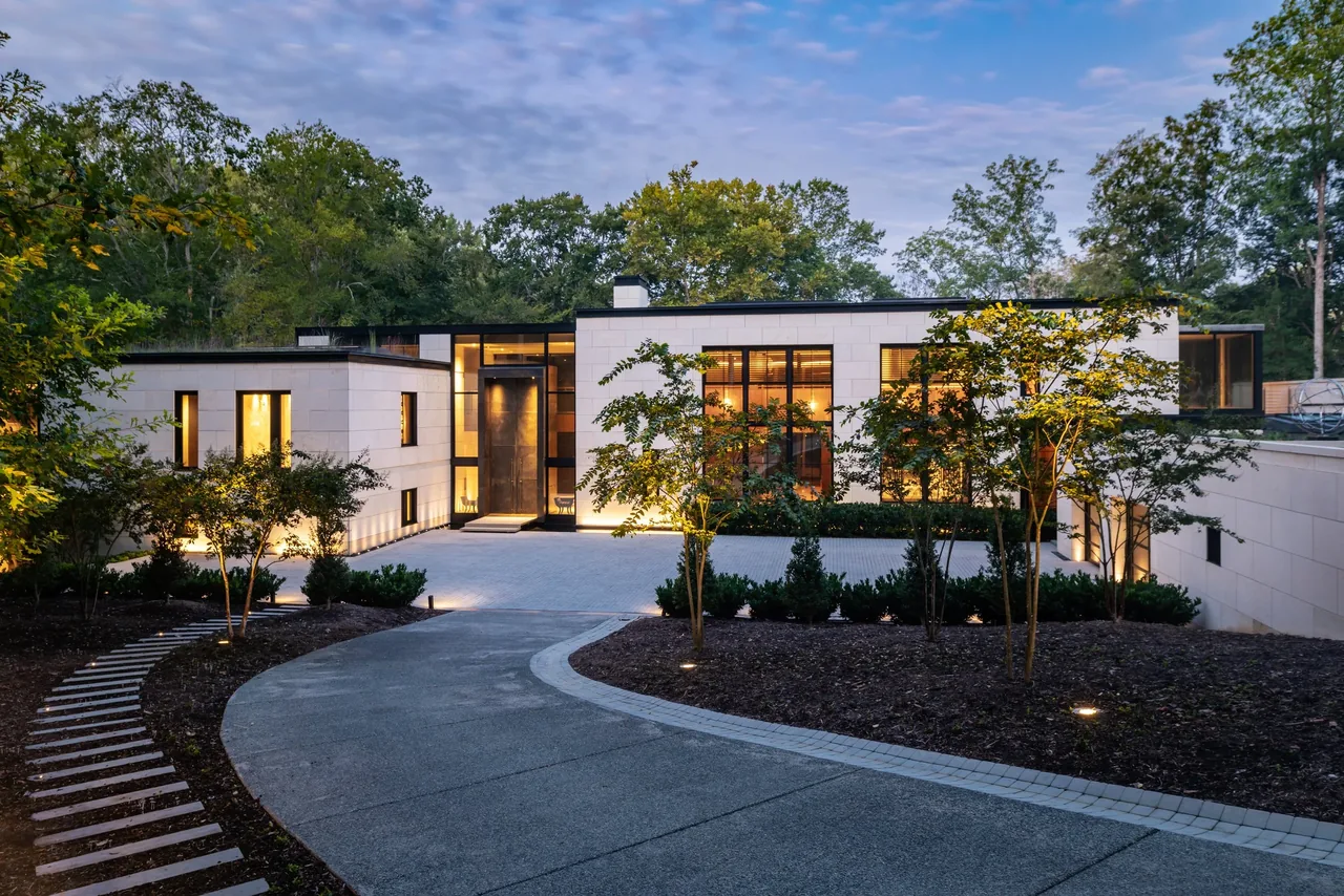 Front exterior view of a modern single-story house with large windows and illuminated landscaping at dusk.