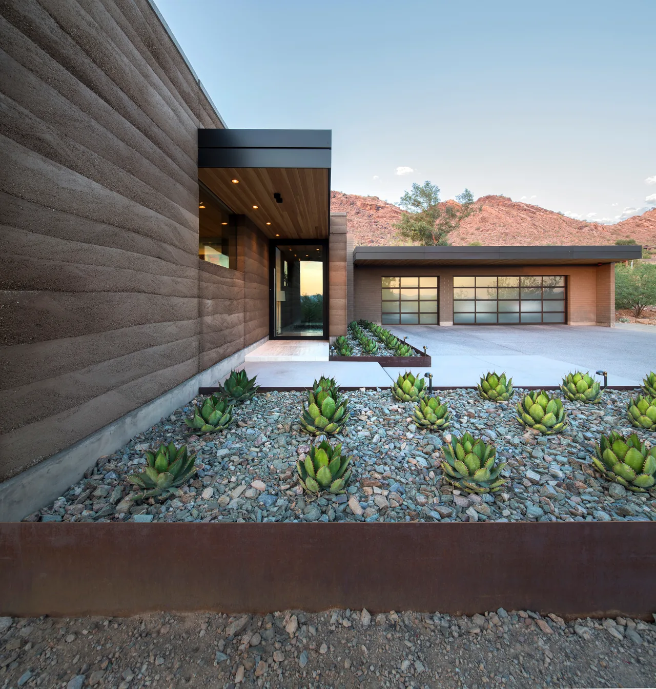 Entryway and garage of a modern home with rammed earth walls and desert landscaping with succulents.