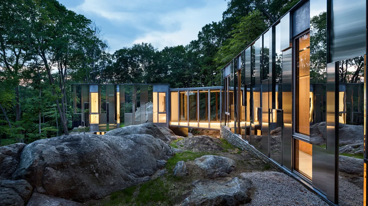 Modern house with reflective glass exterior walls connected by a glass-enclosed bridge over rocky terrain at dusk.