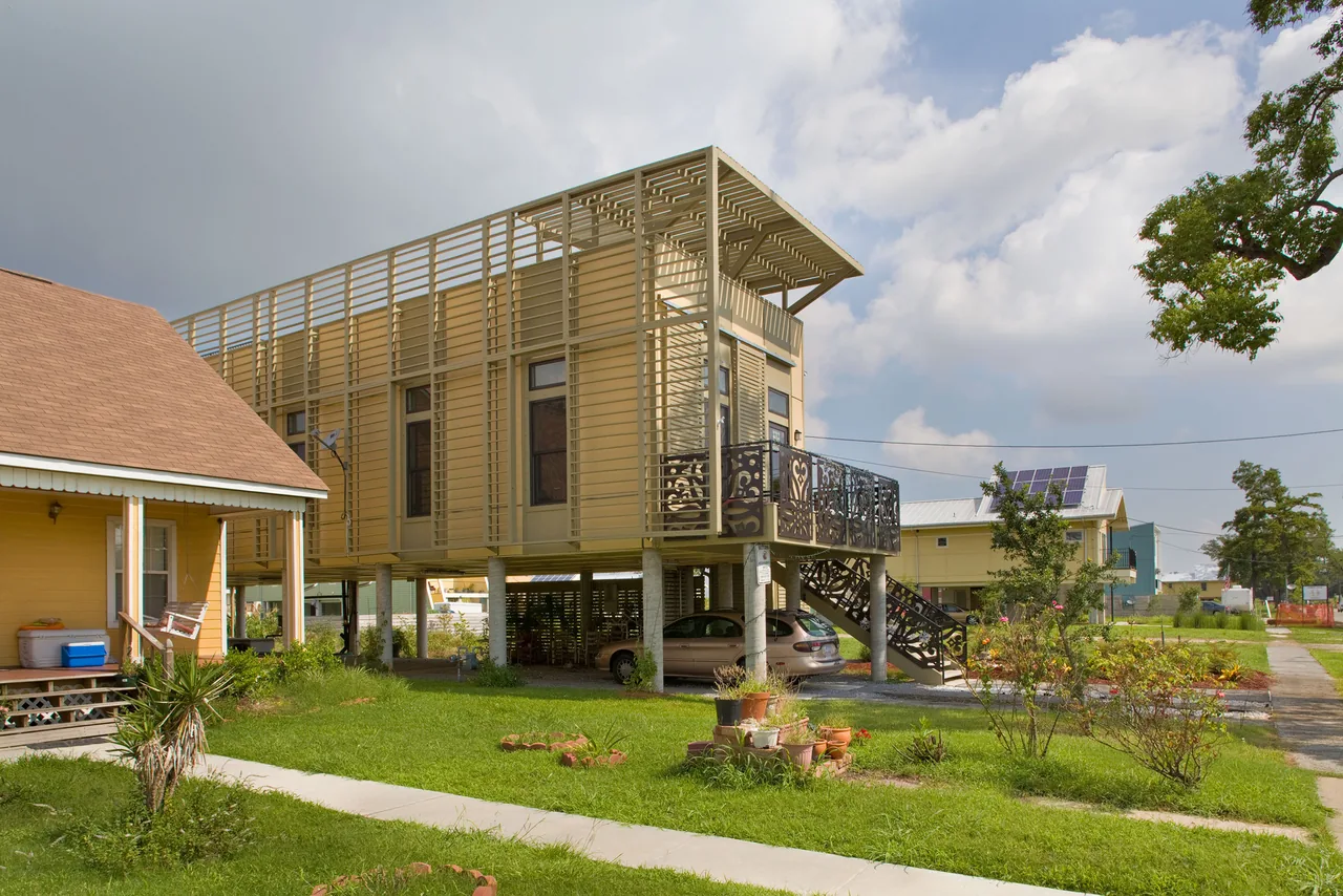 Elevated modern house with beige horizontal slats and decorative metal balcony railing on stilts over garage.