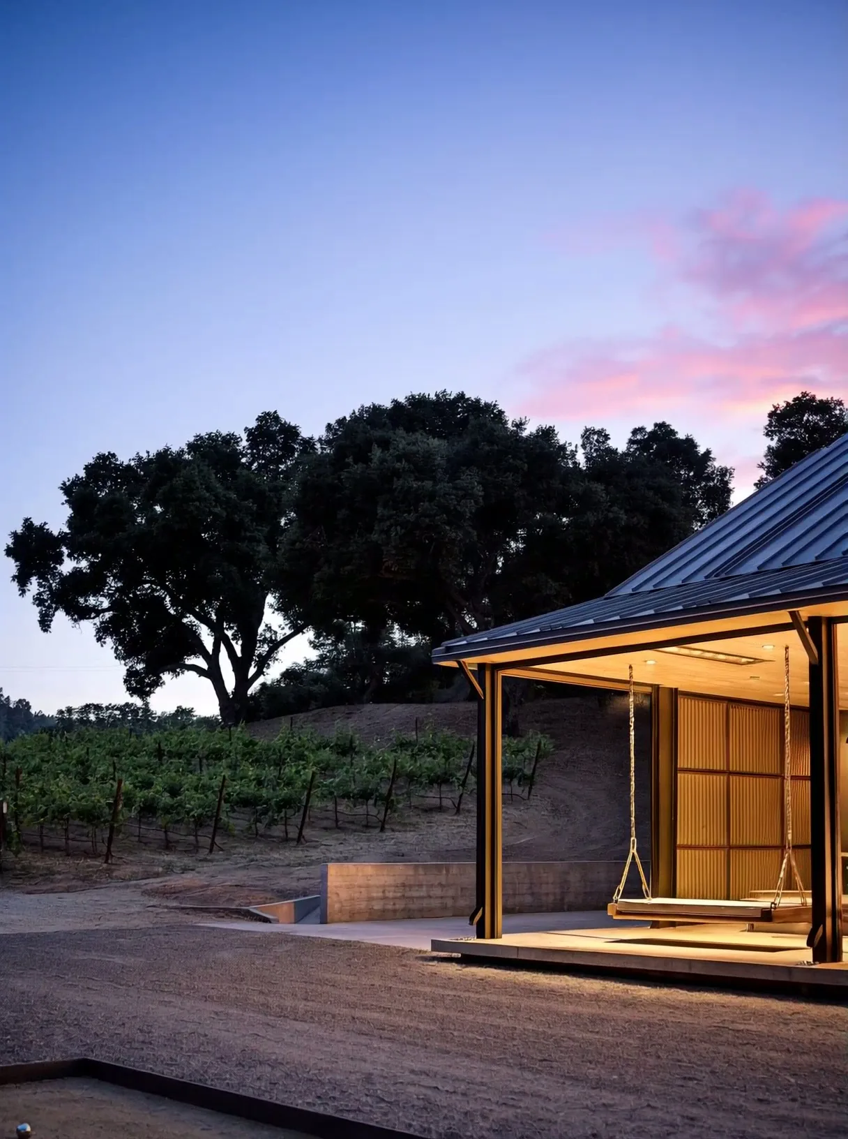 Open pavilion with two wooden swings hanging under a metal roof at dusk near a vineyard and oak trees.