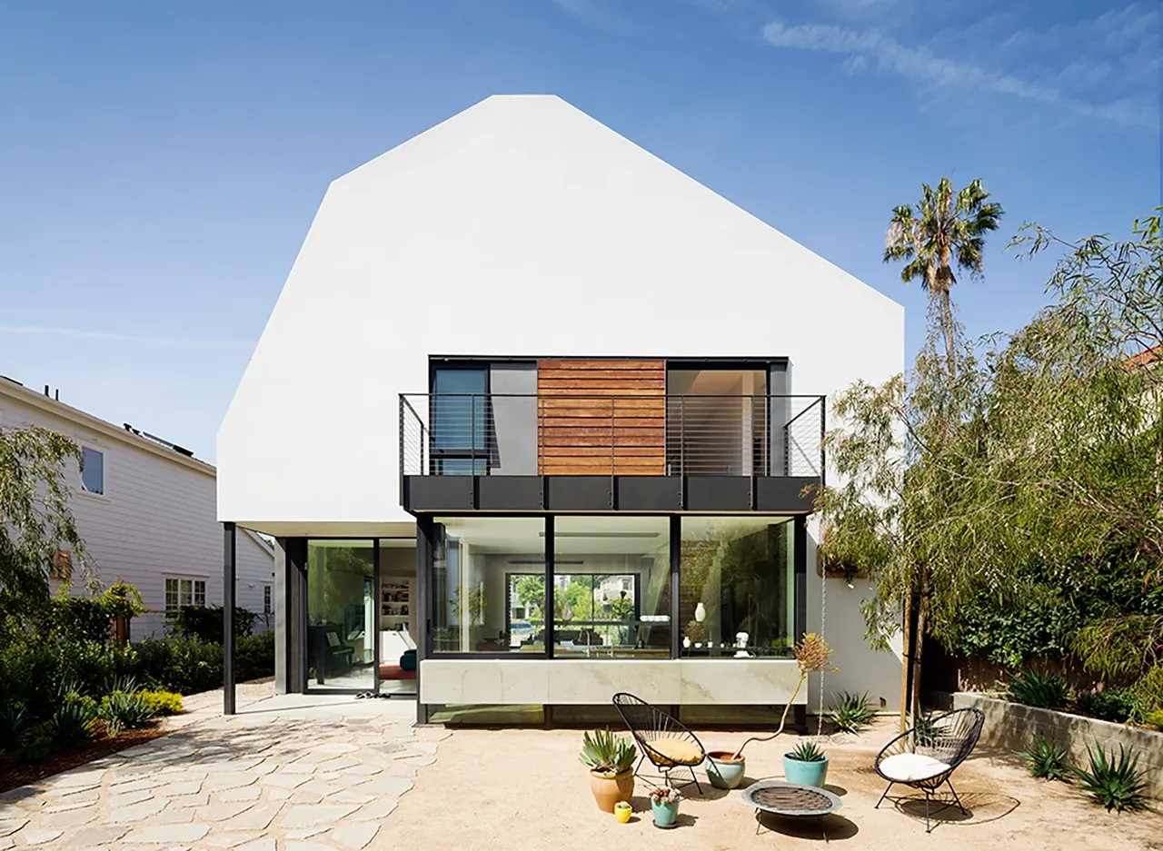 Modern white two-story house with large glass windows and a balcony overlooking a sandy backyard with chairs and potted plants.