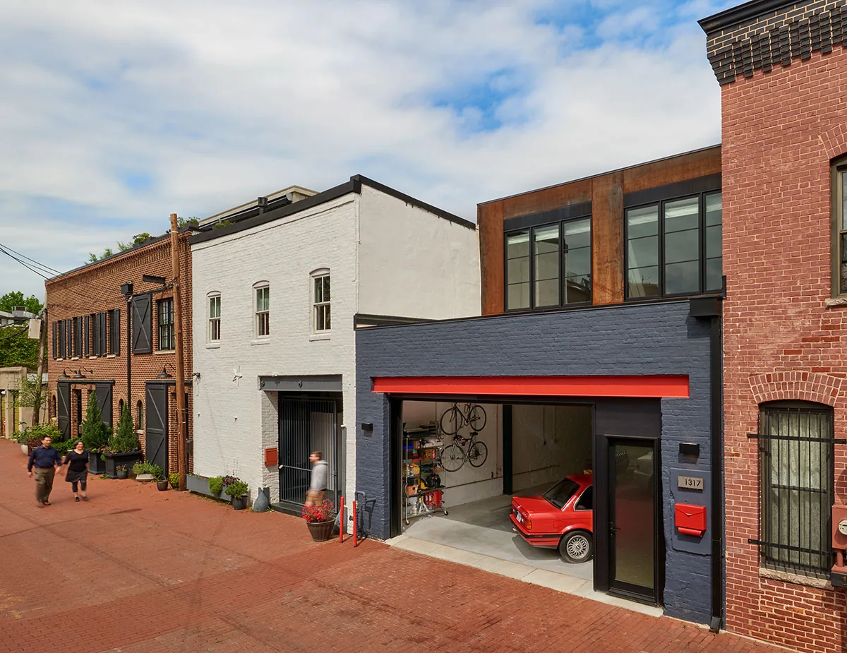 Street view of a modern garage with dark blue brick walls and large black-framed windows above, housing a red car and bicycles.