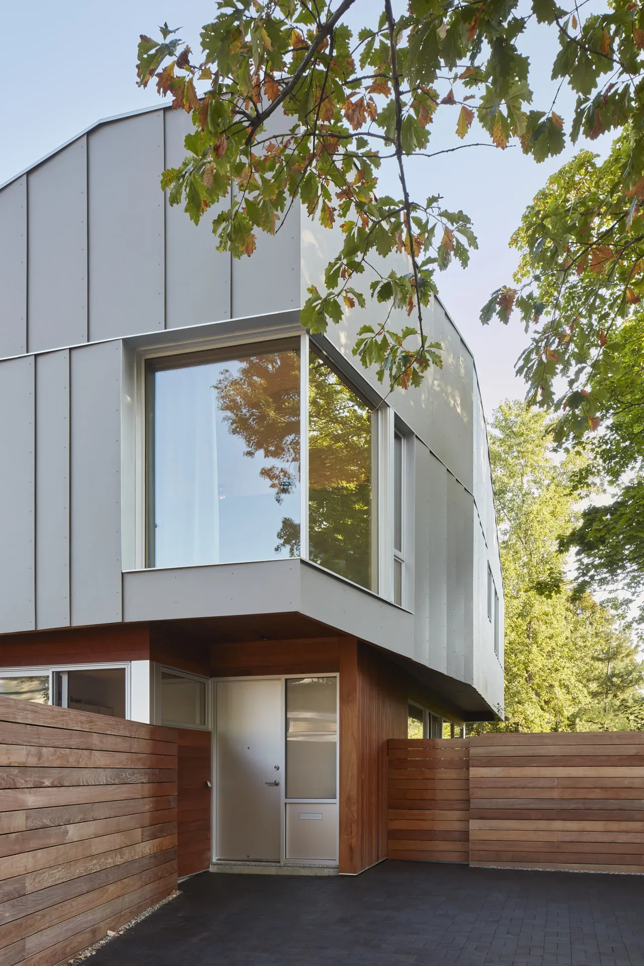 Modern two-story house exterior with metal cladding, a large corner window, wooden fencing, and entry door under an overhang.