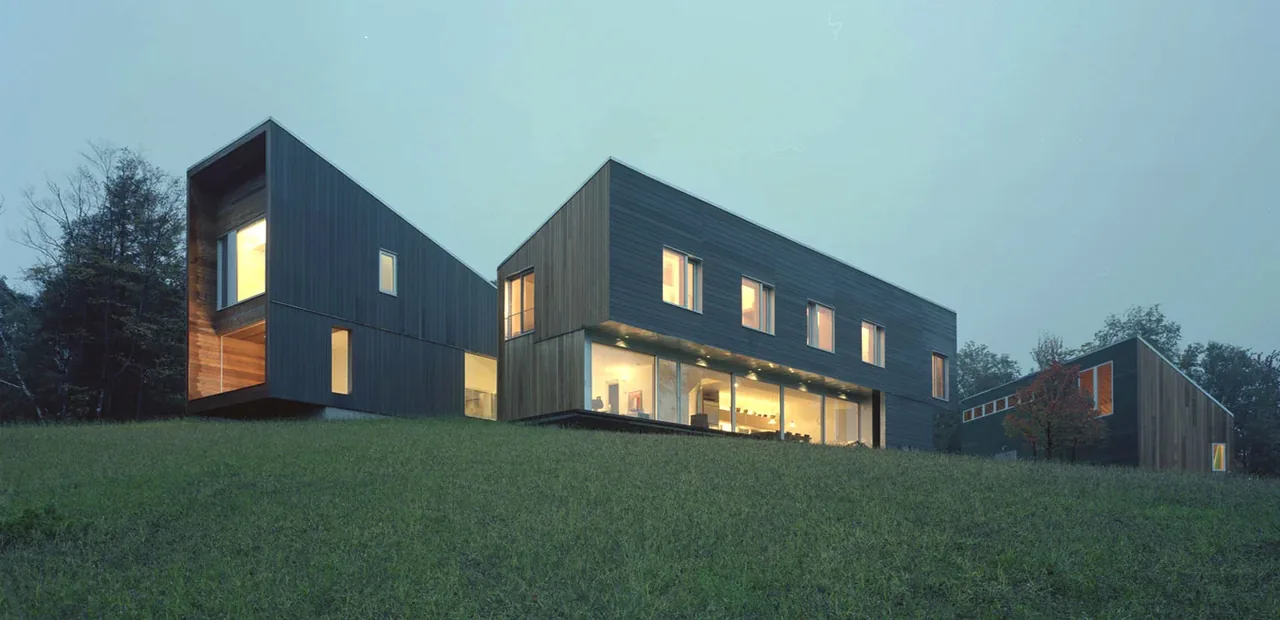 Evening view of modern mountain house with dark wood siding and large windows on grassy hill.