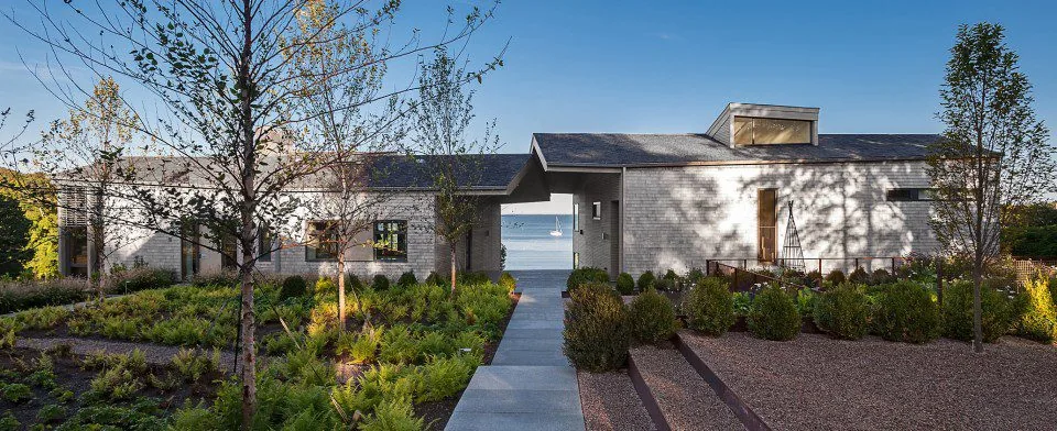 Exterior view of a modern house with shingle siding and a walkway leading to water visible between two wings.