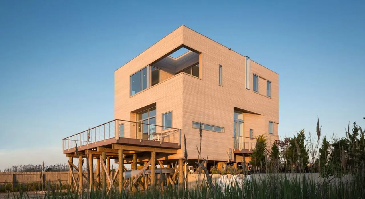 Wooden cube-shaped house elevated on stilts with deck surrounded by vegetation under clear sky.