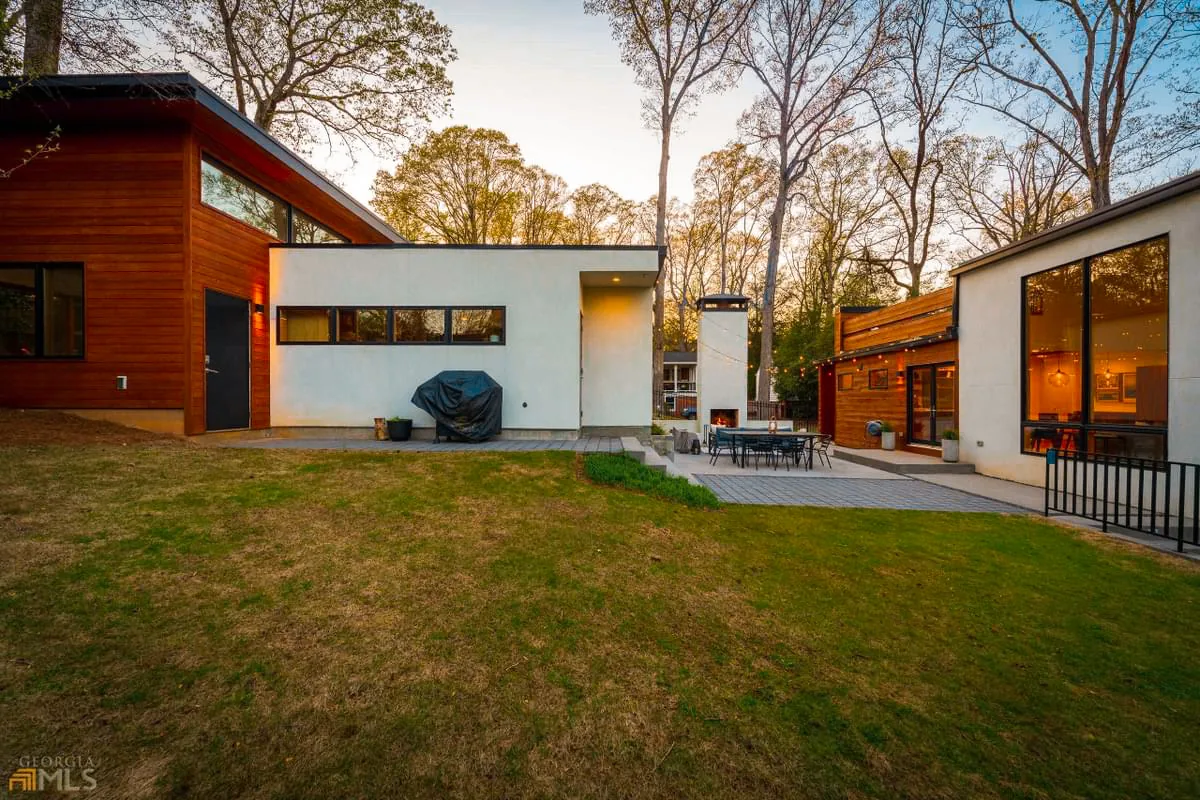 Outdoor patio area of a modern house with a fireplace, surrounded by wood and white stucco exterior walls at dusk.