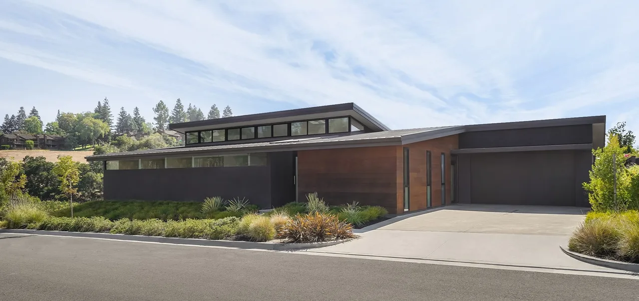 Single-story modern home with flat and shed roofs, dark exterior walls, and wood paneling under a partly cloudy sky.