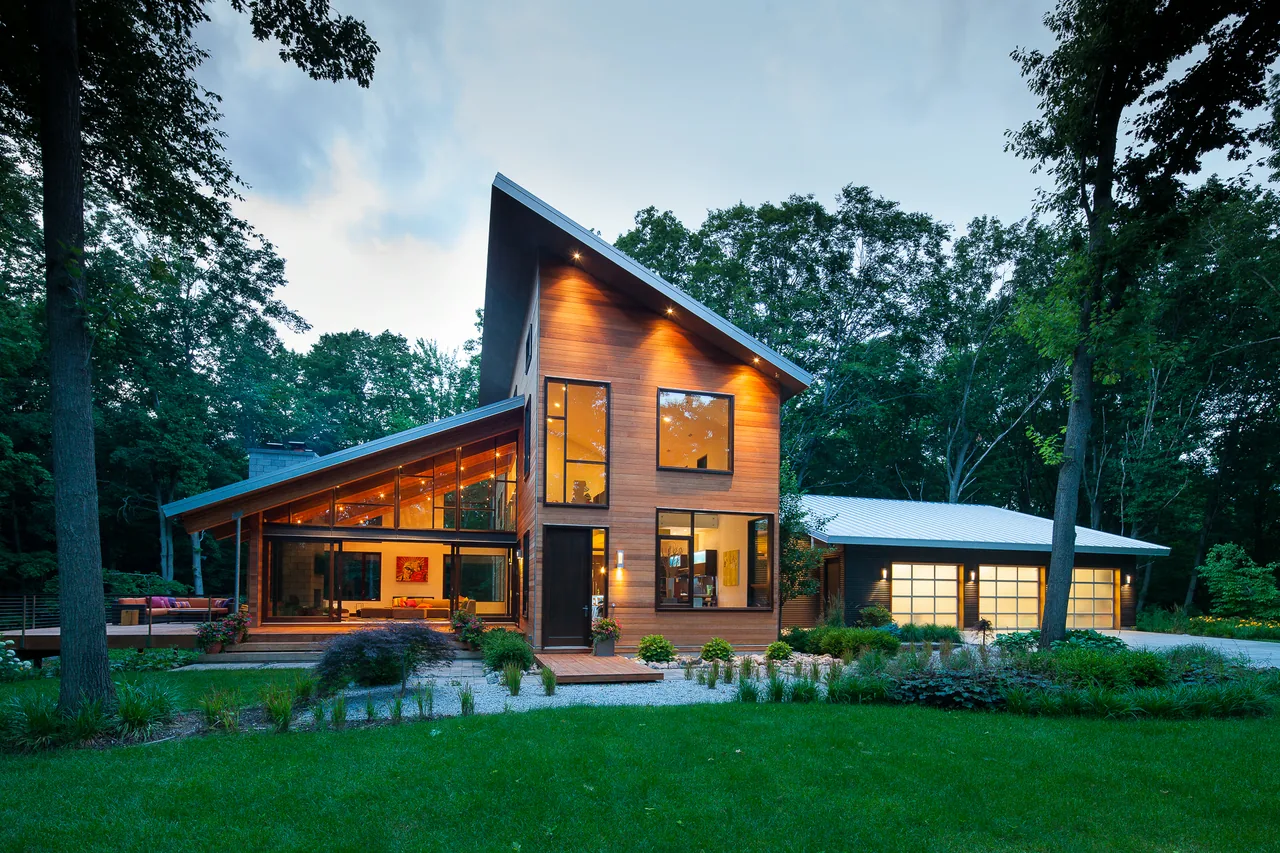 Modern house exterior with sloped roofing, large black-framed windows, and wood siding surrounded by trees at dusk.