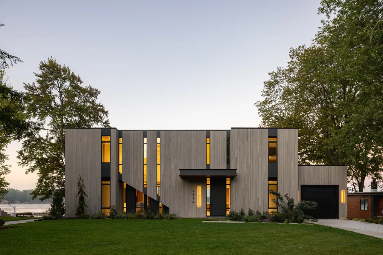 Modern house exterior with vertical light wood siding and large black-framed windows at dusk.