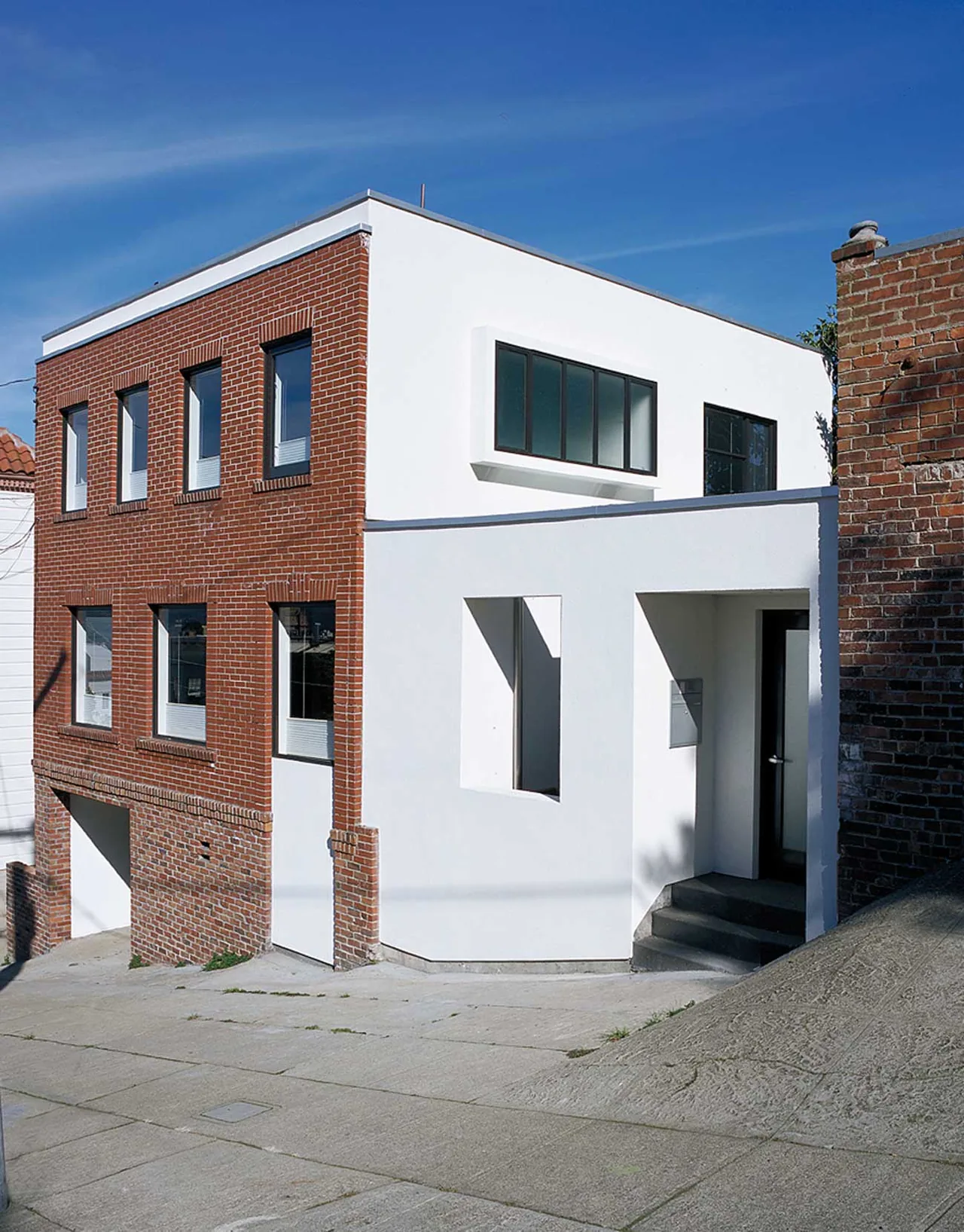 Two-story residential building with a red brick facade on one side and white stucco on the other, featuring rectangular windows and a sloped