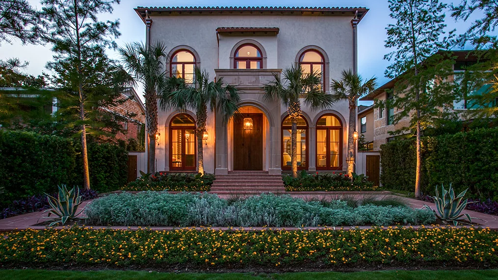 Front view of a two-story Spanish Colonial Revival house with arched windows and door, surrounded by palm trees and landscaping at dusk.