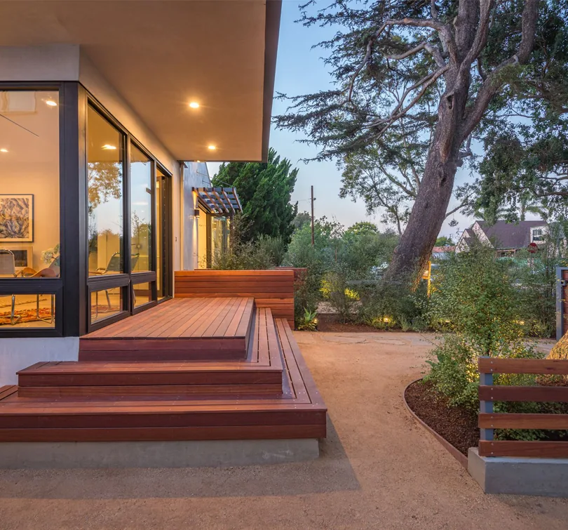 Modern bungalow exterior with a wooden deck and large black-framed windows overlooking a garden area at dusk.