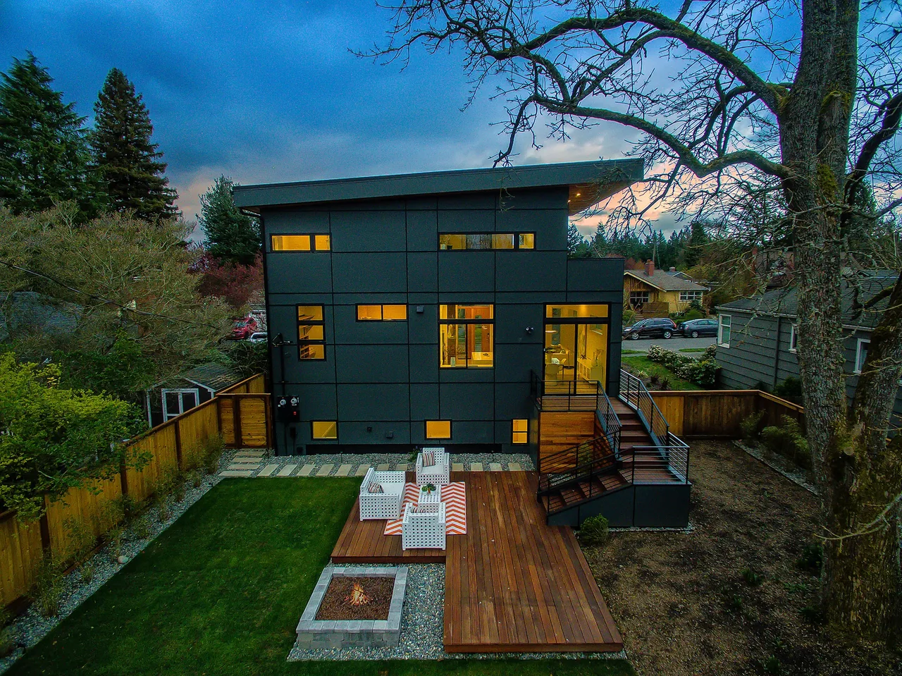 Rear view of a modern two-story house at dusk with large windows, a wooden deck, outdoor seating, and a fire pit in a fenced backyard.