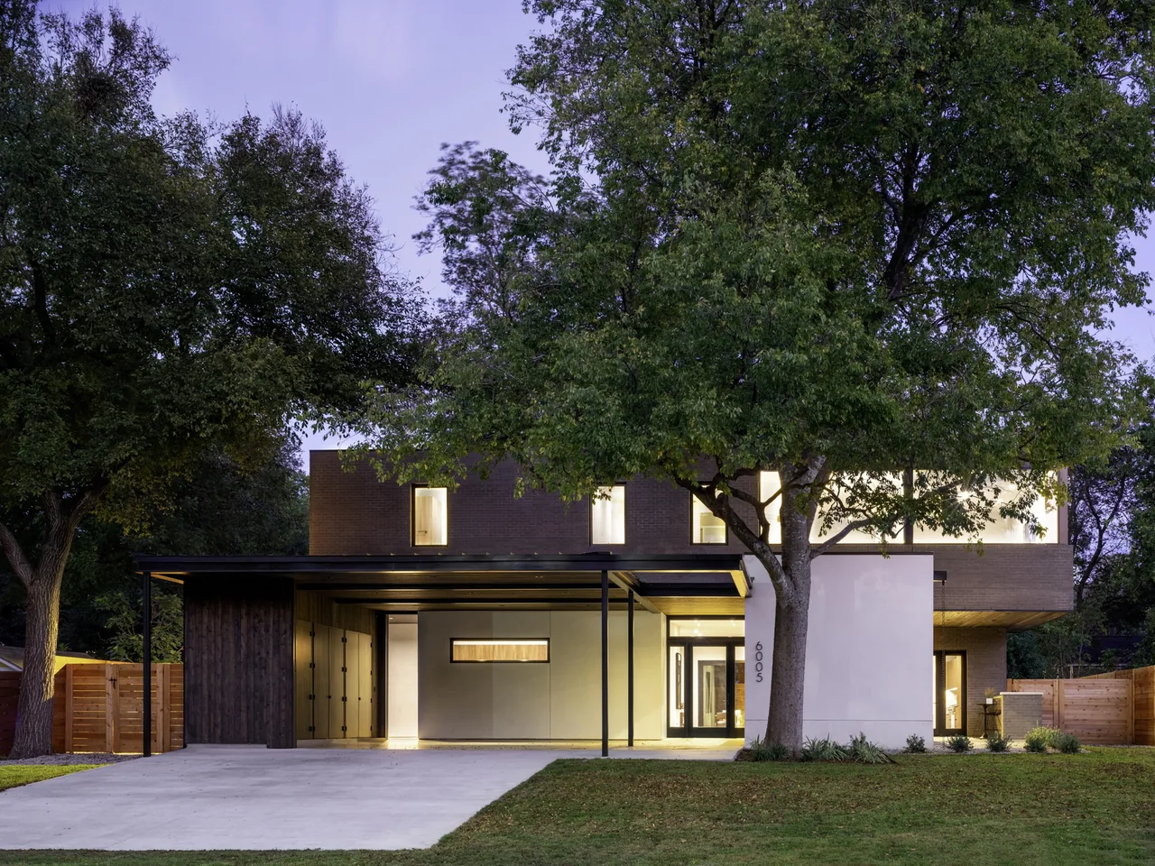 Modern two-story house with flat roof and large windows, surrounded by trees at twilight.