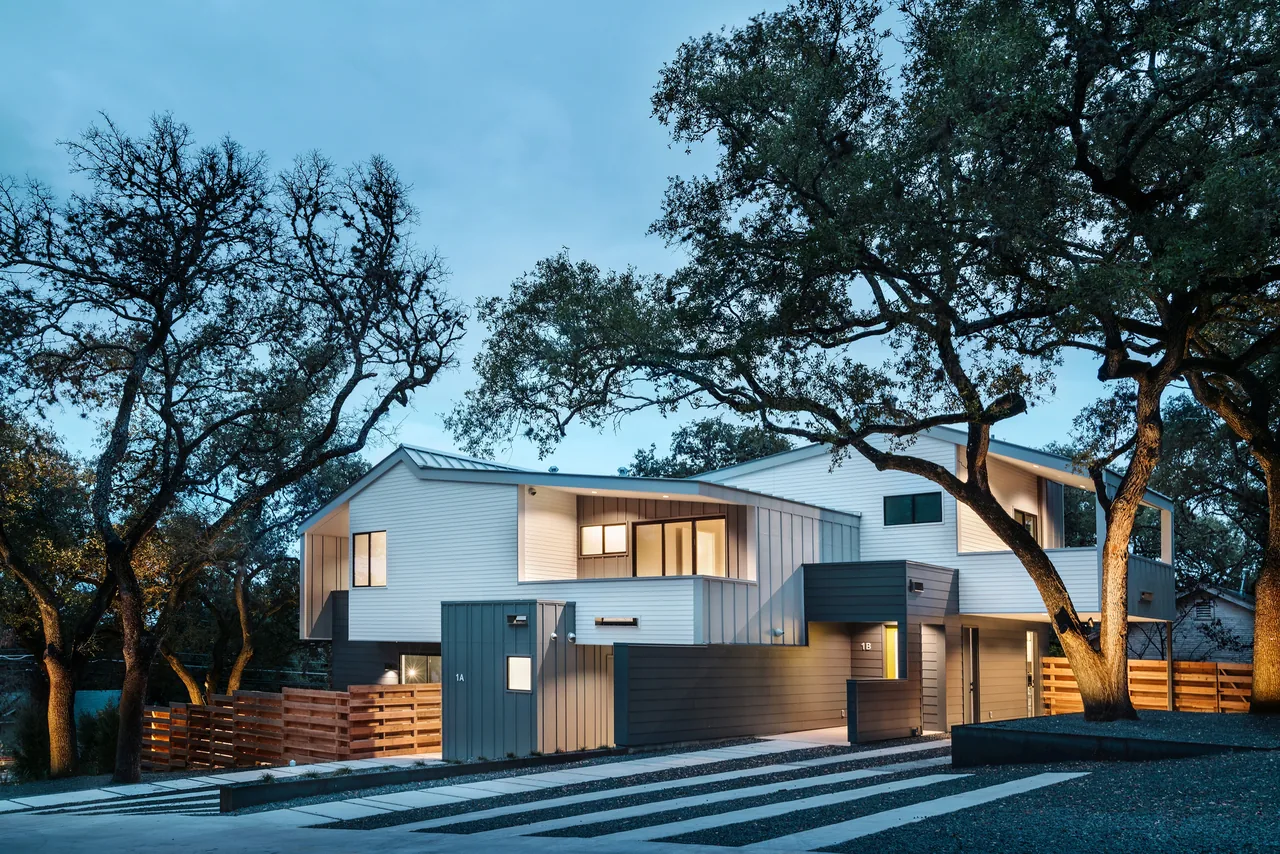Evening view of two modern side-by-side duplexes with white siding and gray accents among trees.